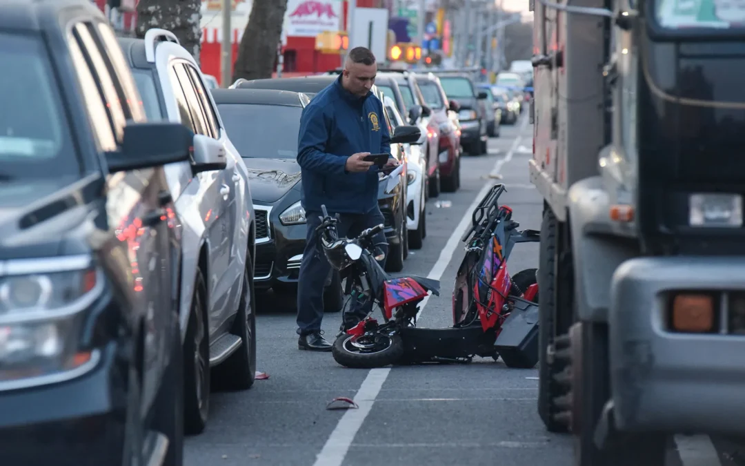 NYPD officer photographing a wrecked red e-scooter in a Brooklyn traffic lane after a crash